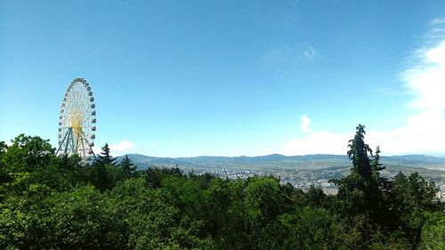 Ferris wheel by trees on mountain against blue sky