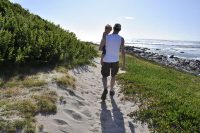 Rear view of man carrying son while walking on sand at beach