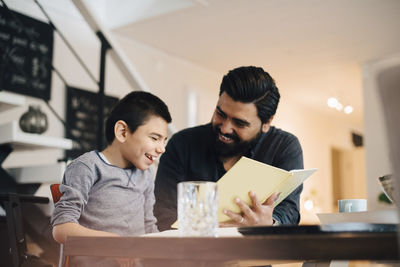 Smiling father teaching autistic son while sitting at table in house