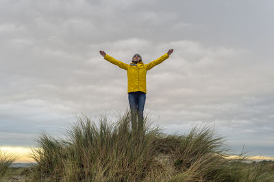 Woman wearing a yellow coat on top of a sand dune with her arms raised, success concept.