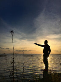 A silhouette of a man standing in sunset in front of a lake aiming a gun