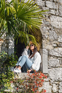 Attractive young woman in spring outfit sitting in window of stone building in park.