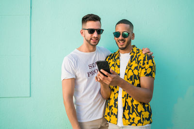 Young man using mobile phone by gay friend while standing against wall