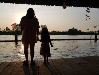 Rear view of women standing by lake against sky during sunset