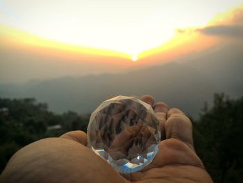 Cropped hand holding crystal ball against sky during sunset