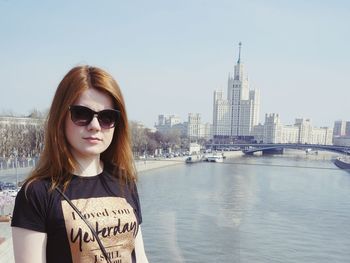 Portrait of young woman standing in city by river against sky
