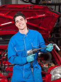 Portrait of smiling mechanic with equipment standing against car in garage