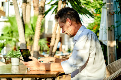 Side view of middle aged businessman in smart casual clothes scrolling data on cellphone while sitting at table with laptop during work in street restaurant