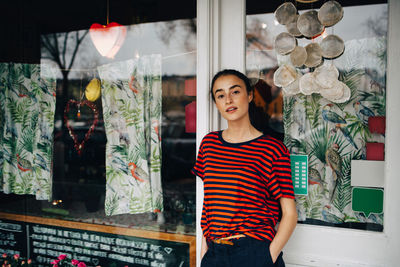 Portrait of confident young woman standing with hand in pocket against restaurant door