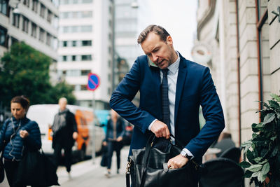 Mature businessman talking smart phone while searching in bag on sidewalk in city