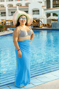 A smiling brunette woman in a swimsuit and a hat stands by the pool. vertical photo