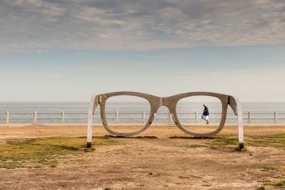 Senior man walking on beach seen through eyeglasses sculpture against sky