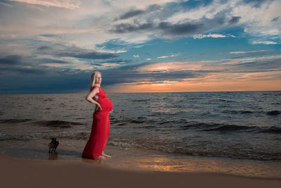 Portrait of pregnant woman standing on shore at beach against cloudy sky