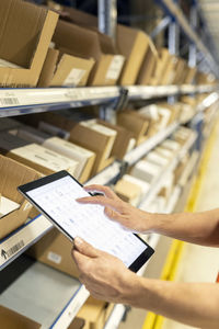 Hands of worker analyzing list on tablet pc by rack in warehouse