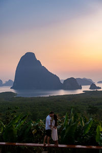 Rear view of man and woman standing on mountain against sky during sunset