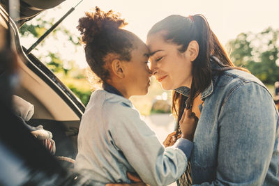 Smiling woman and daughter sitting face to face with eyes closed in car trunk during picnic