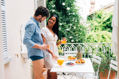 Smiling couple with food and drink on table standing in balcony