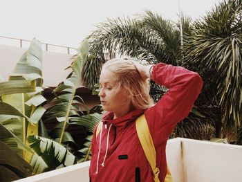 Mid adult woman with hand in hair standing by wall against trees