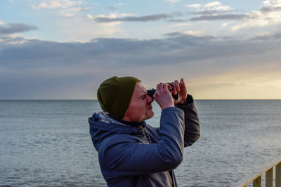 Profile view of young man looking through binoculars against sea during sunset