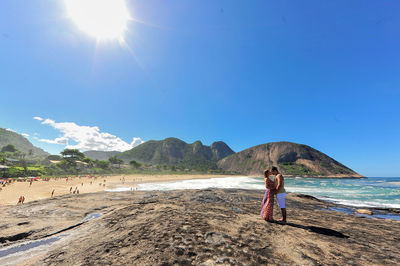 Side view of couple kissing at rocky beach against clear sky on sunny day