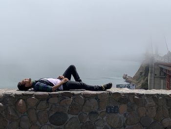 Full length of man lying on retaining wall against sky