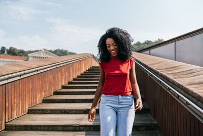 Smiling young african american woman in red t shirt and jeans walking down wooden stairway at city street during sunny day