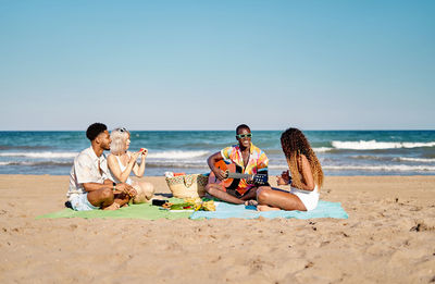 Happy multiethnic man and women in casual clothes smiling and eating fruits while sitting cross legged on blanket and listening to black friend playing acoustic guitar near sea on summer weekend day on beach