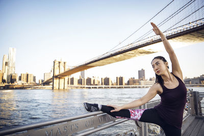 Female athlete stretching leg and arm on promenade with brooklyn bridge in background