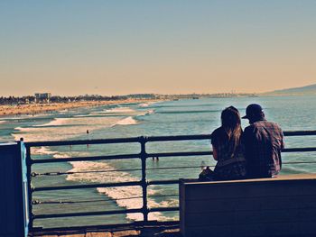 Rear view of couple sitting on pier by sea against sky