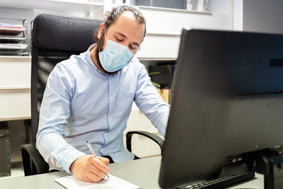 Focused young bearded male manager in formal blue shirt and protective mask making notes in notebook while sitting in chair in front of computer monitor in modern workplace