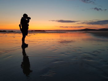 Silhouette man photographing while standing at beach against sky during sunset