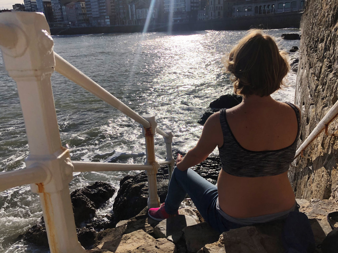 REAR VIEW OF WOMAN LOOKING AT SEA WHILE SITTING ON RAILING