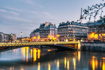 Bridge over river by illuminated buildings against sky at night