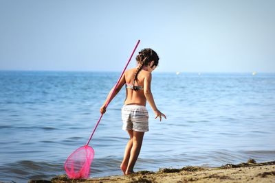 Rear view of girl in swimwear holding fishing net while walking on beach against clear sky