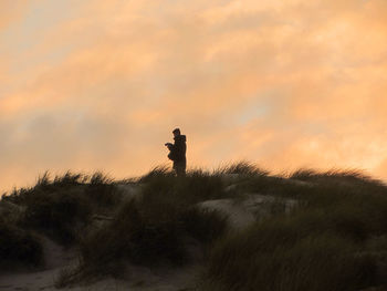 Low angle view of man standing on sand dune with bushes against sky during sunset