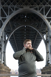 Overweight man holding headphones while standing below williamsburg bridge in city