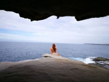 Woman sitting on beach cliff by sea against sky