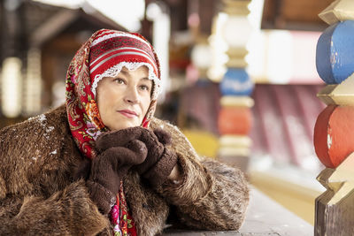 A slavic woman in a national colored scarf, a fur coat and  mittens on the porch of a wooden house