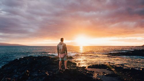 Rear view of man standing on beach against sky during sunset