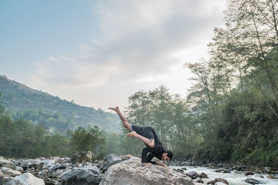 Man doing yoga on rock at riverbank in forest