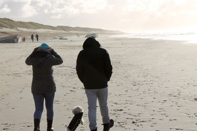 Rear view of silhouette couple with dog walking at beach against sky during sunny day