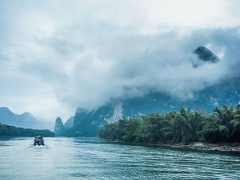 Boat sailing in river by mountains against cloudy sky