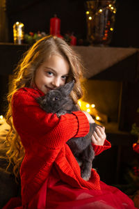 A little blonde girl with a gray rabbit in her arms next to a christmas tree decorated with garlands