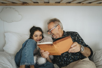 Happy senior man reading storybook with granddaughter on bed at home