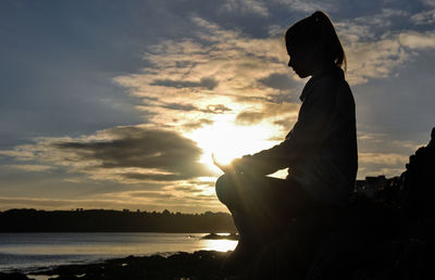 Silhouette woman doing yoga at lake against sky during sunset