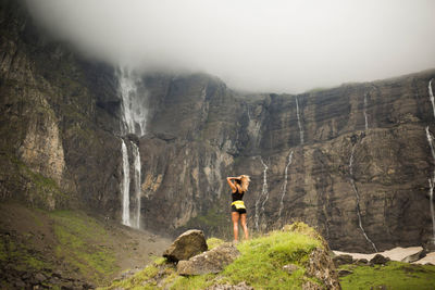 Low angle back view of female tourist standing on stone against cliff of monte perdido with waterfalls on overcast day in pyrenees