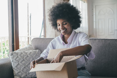 Smiling woman opening box while sitting on sofa at home