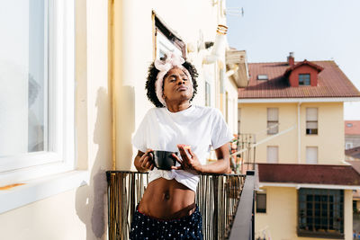 Relaxed african american female with toast standing on balcony and sipping hot beverage in morning