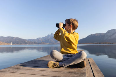 Boy looking through binocular sitting on pier