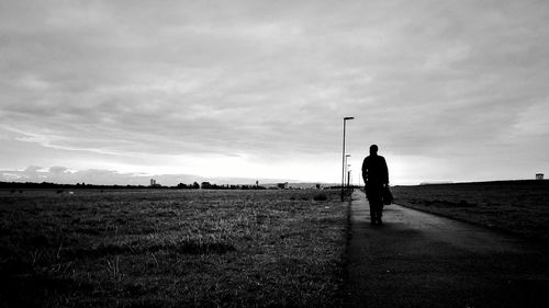 Rear view of silhouette man walking on field against cloudy sky